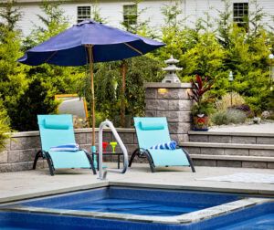 Poolside chairs with an umbrella and garden in the background. Dex by Terra landscape and hardscape design build project in Westminster, Massachusetts.