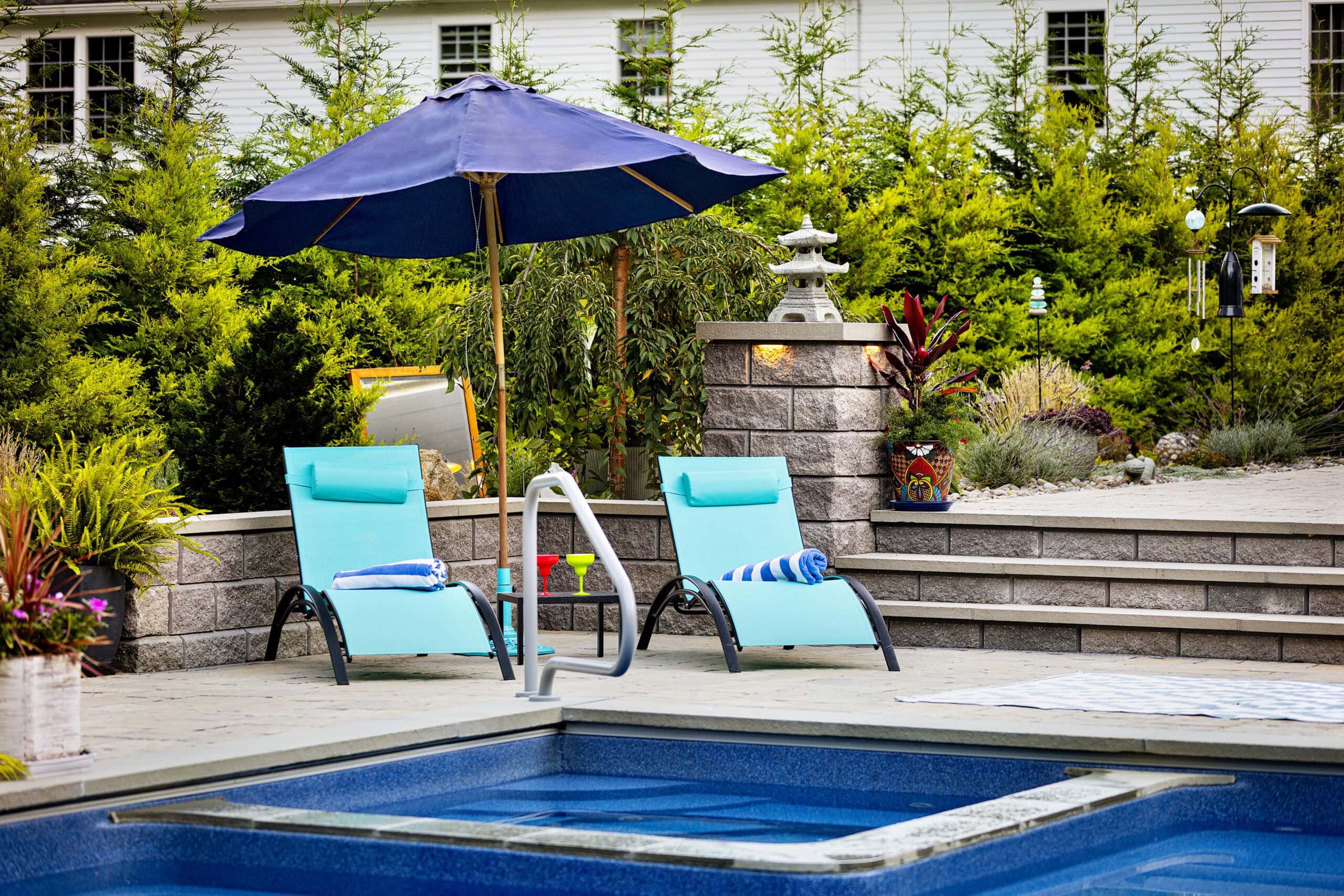 Poolside chairs with an umbrella and garden in the background. Dex by Terra landscape and hardscape design build project in Westminster, Massachusetts.