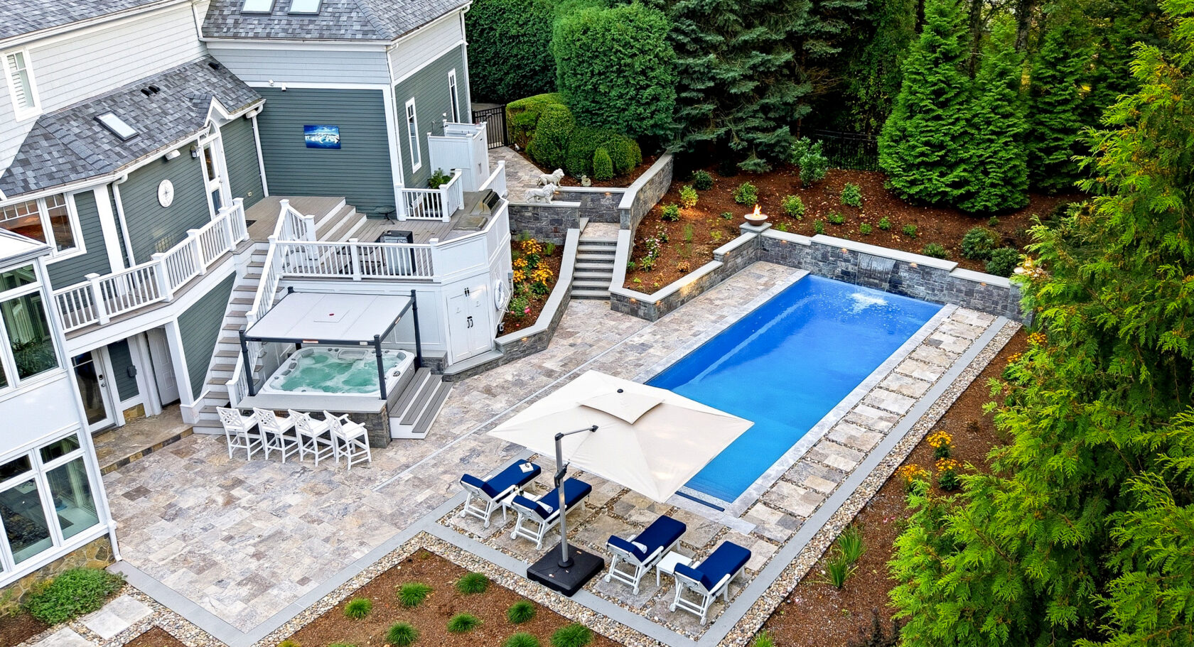 Drone view of pool deck, patio, hot tub bar, and landscaping surrounding the pool. Dex by Terra Hardscaping and Landscaping project in Lincoln, MA.