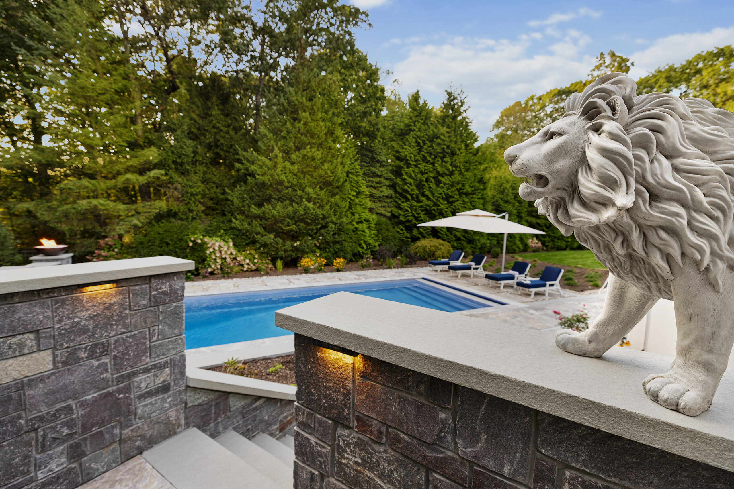 A lion statue guards the stone stairs with the pool in the background. Dex by Terra hardscape project in Lincoln, MA.