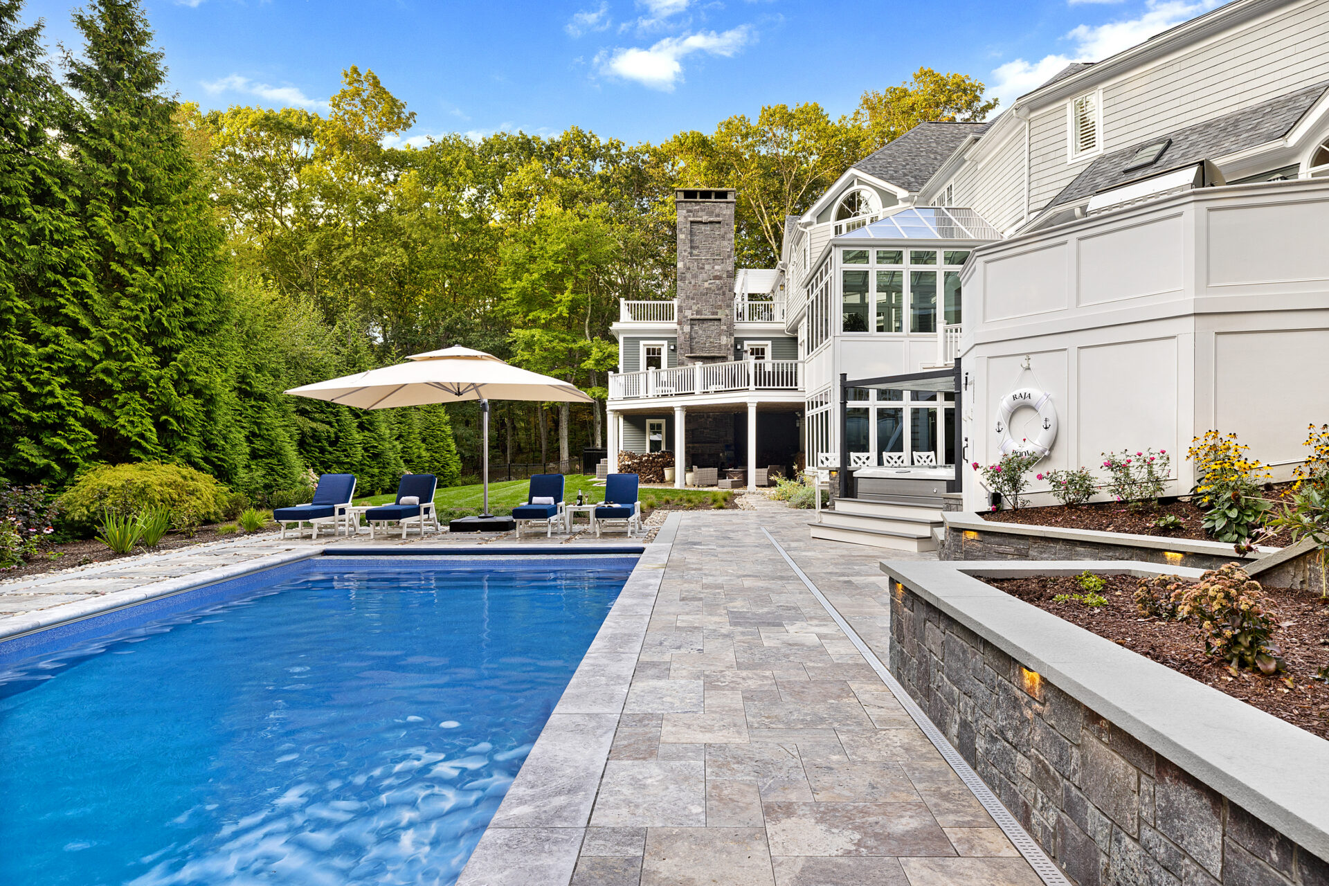View of the pool, pool deck, veneer stone wall, and back of the home. Dex by Terra hardscape project in Lincoln, MA.