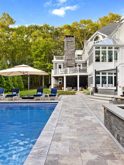 View of the pool, pool deck, veneer stone wall, and back of the home. Dex by Terra hardscape project in Lincoln, MA.