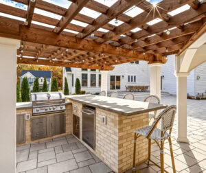 Outdoor kitchen with seating under a patio pergola, all designed and built by Dex by Terra in North Attleboro, MA.