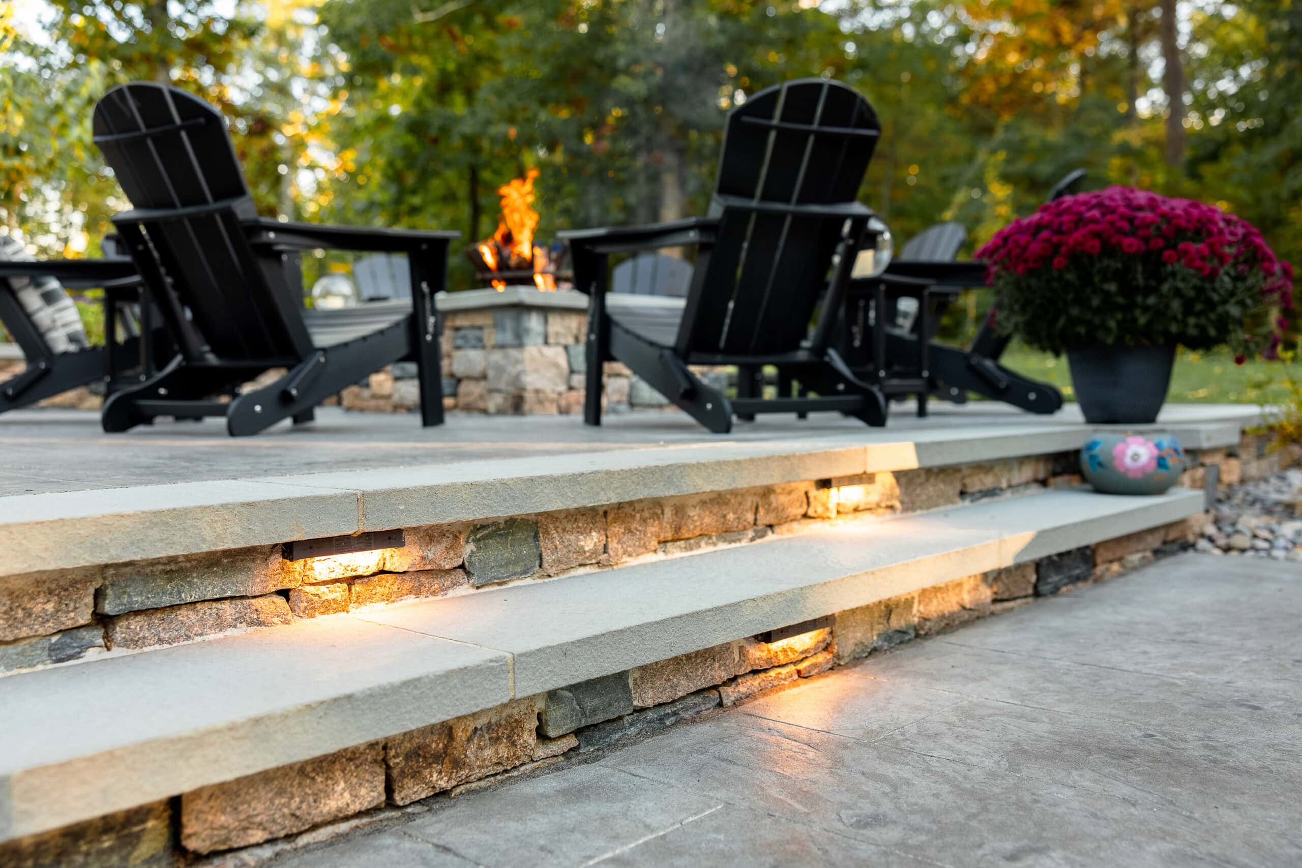 Low voltage LED landscape lighting integrated into the stone veneer steps with fire pit in the background. Landscape design in Dunstable, MA built by Dex by Terra.