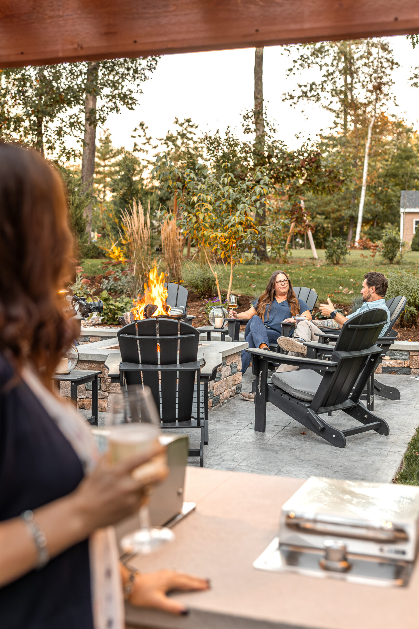 The homeowner watches friends gathered around the fire pit, from the outdoor kitchen area. Landscape design in Dunstable, MA built by Dex by Terra.