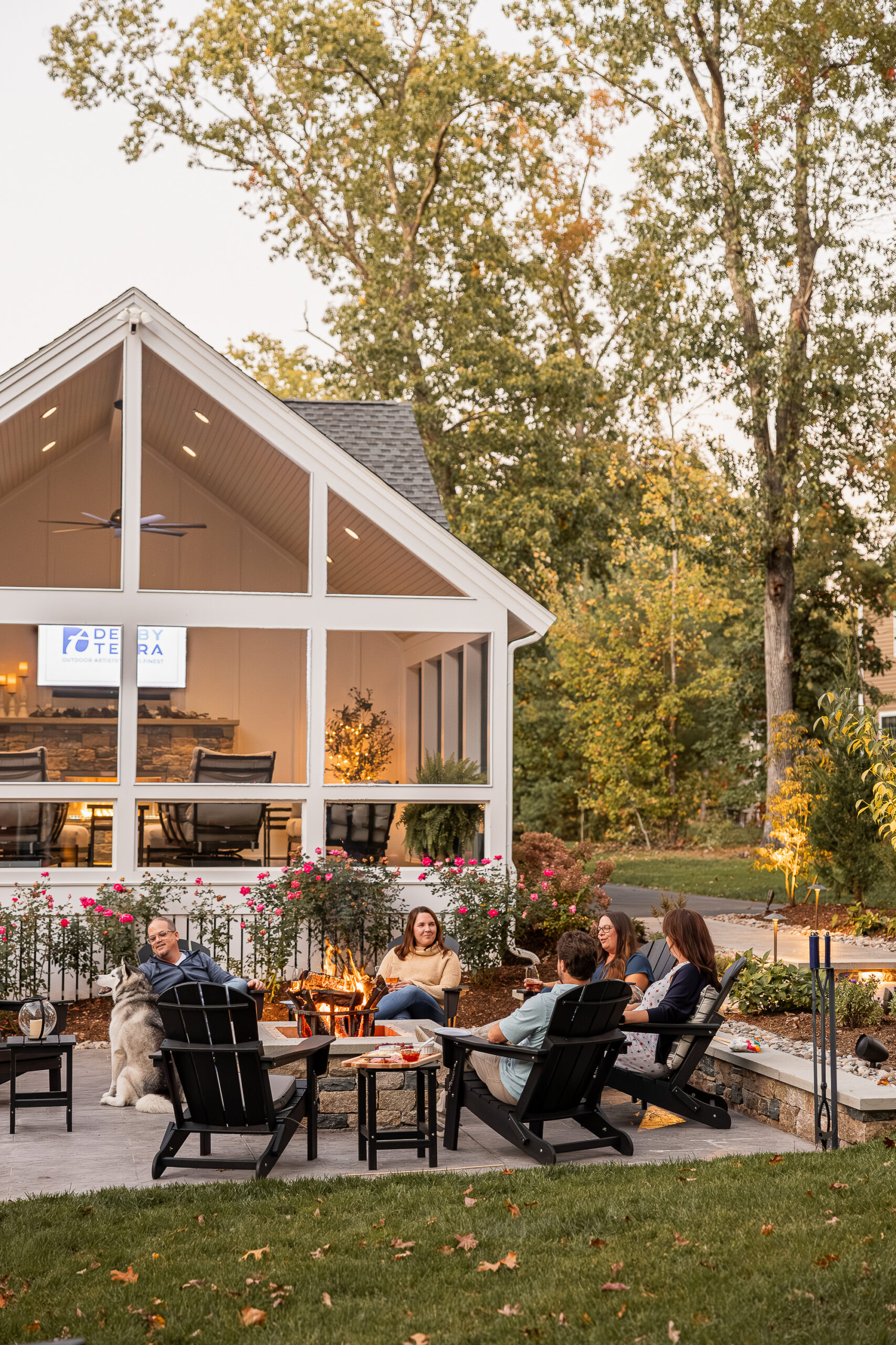 Friends enjoy the fire pit with the screened-in porch in the background as evening falls. Landscape design in Dunstable, MA built by Dex by Terra.