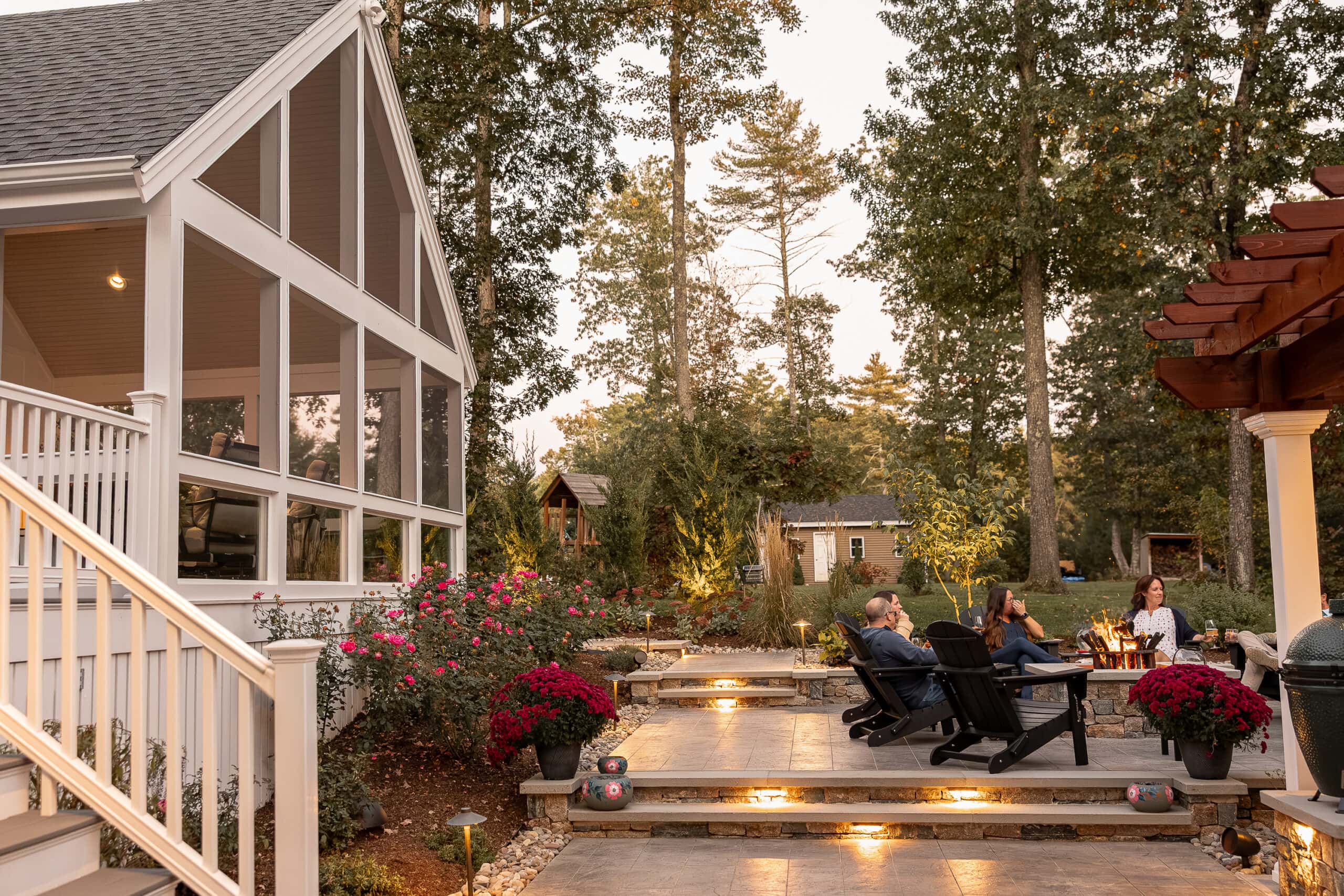 Friends enjoy the fire pit with the screened-in porch in the background as evening falls. Landscape design in Dunstable, MA built by Dex by Terra.
