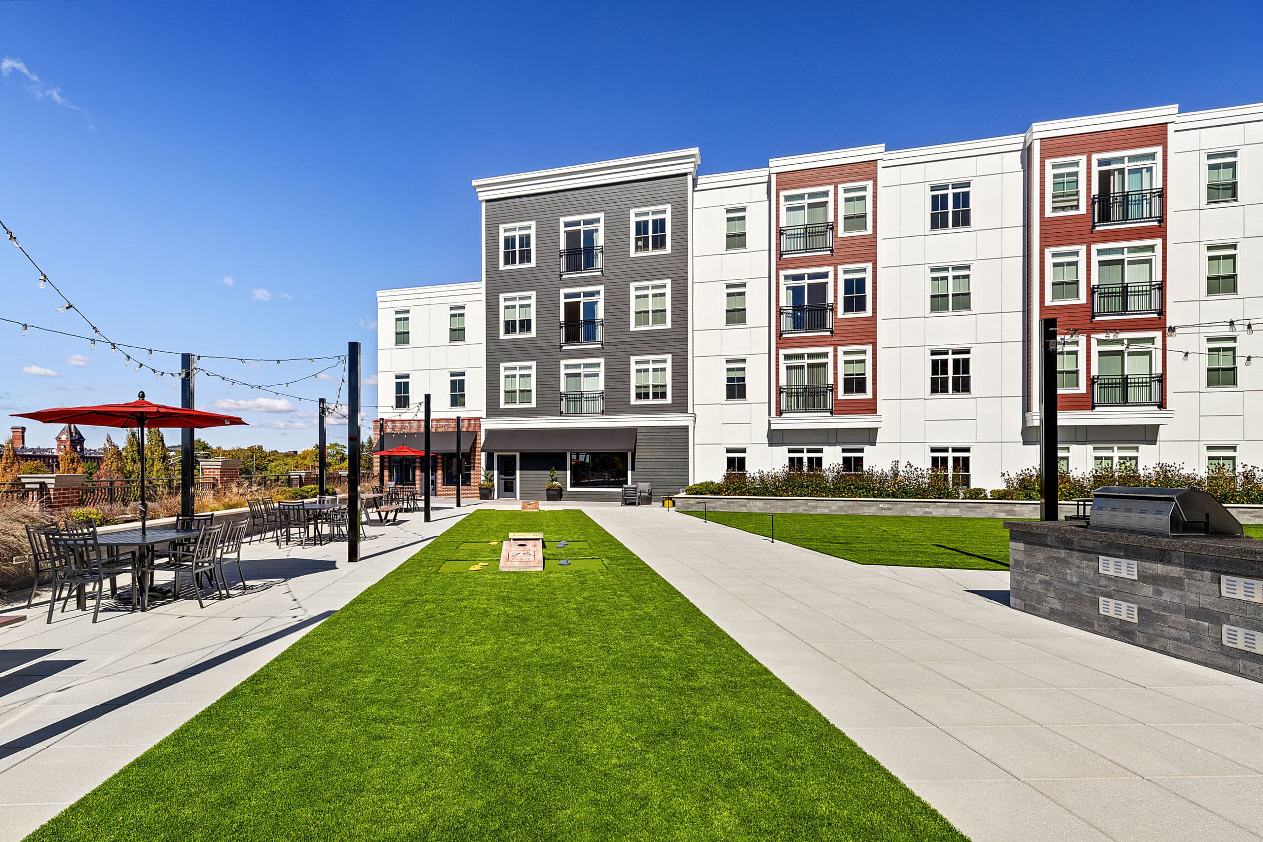 The second floor courtyard and amenity area with tables, chairs, and cornhole areas at 75 Canal Apartments in Manchester, NH. Hardscaping and landscaping by Dex by Terra of Hudson, MA.