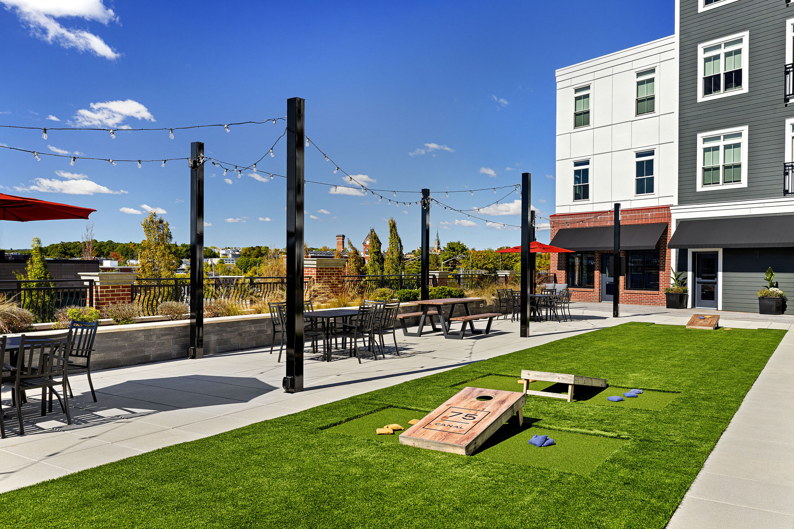 The second floor courtyard and amenity area with tables, chairs, and cornhole areas at 75 Canal Apartments in Manchester, NH. Hardscaping and landscaping by Dex by Terra of Hudson, MA.
