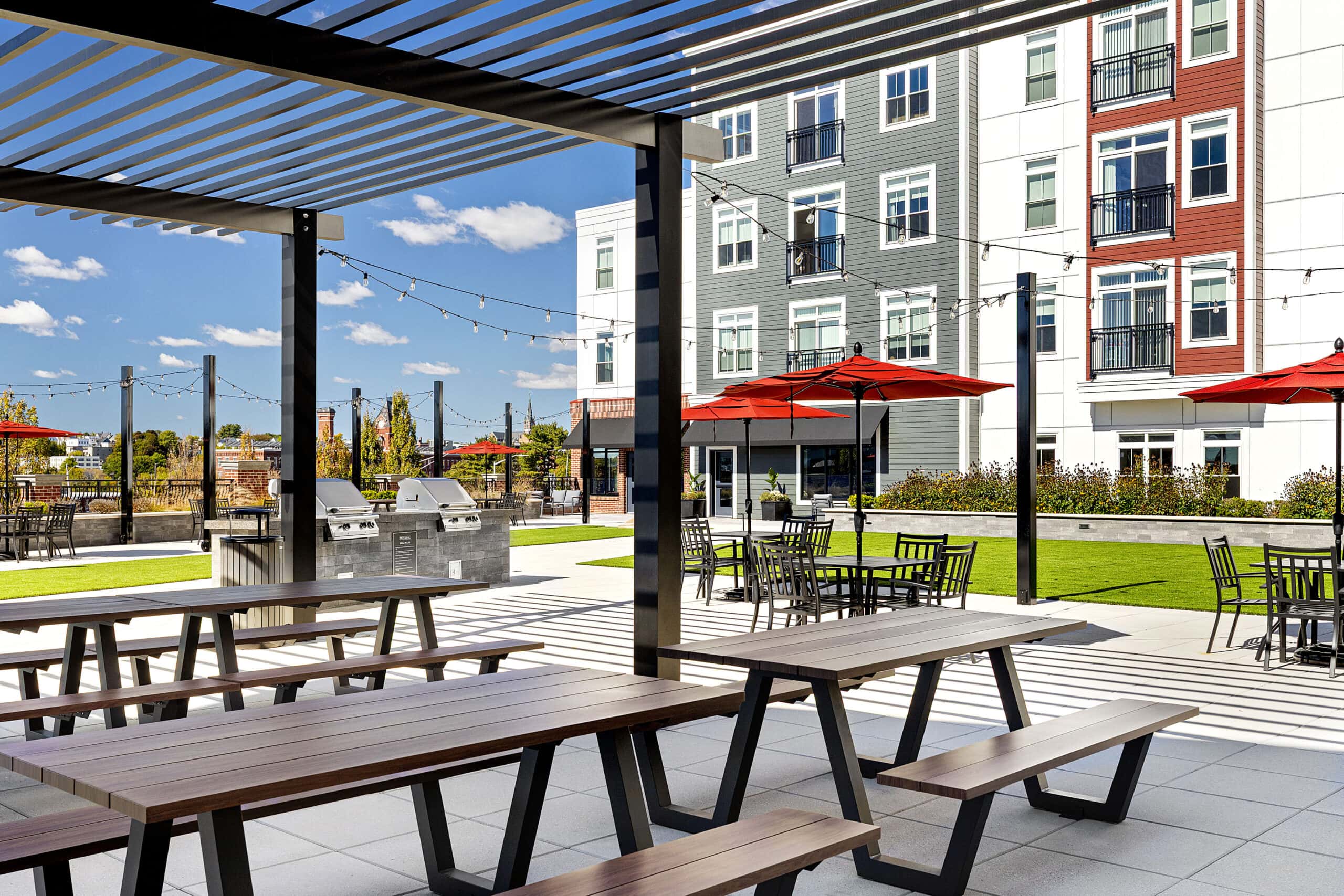 The second floor courtyard amenity area with picnic tables shaded by pergolas. Artificial turf in the background. 75 Canal Apartments in Manchester, NH. Hardscaping and landscaping by Dex by Terra of Hudson, MA.
