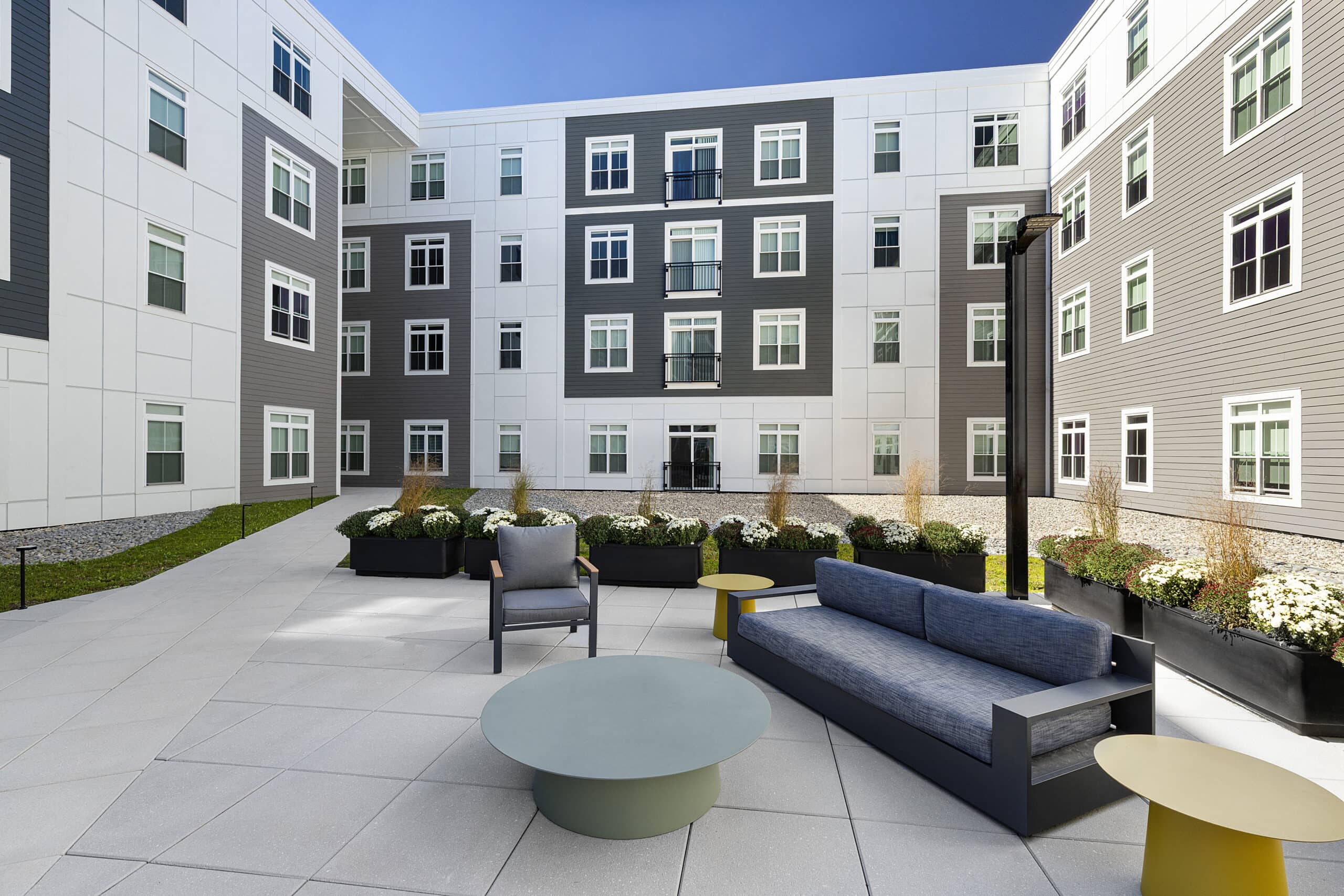 A concrete paver patio with seating, tables and planters in the smaller second floor courtyard area at 75 Canal Apartments in Manchester, NH. Hardscaping and landscaping by Dex by Terra of Hudson, MA.