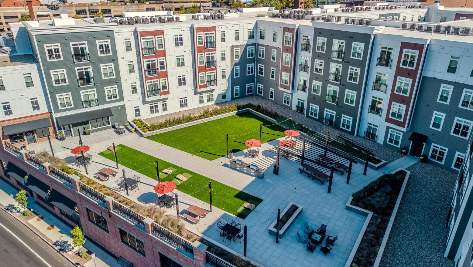 Drone view of the second floor courtyard amenity area with picnic tables shaded by pergolas. Artificial turf in the background. 75 Canal Apartments in Manchester, NH. Hardscaping and landscaping by Dex by Terra of Hudson, MA.