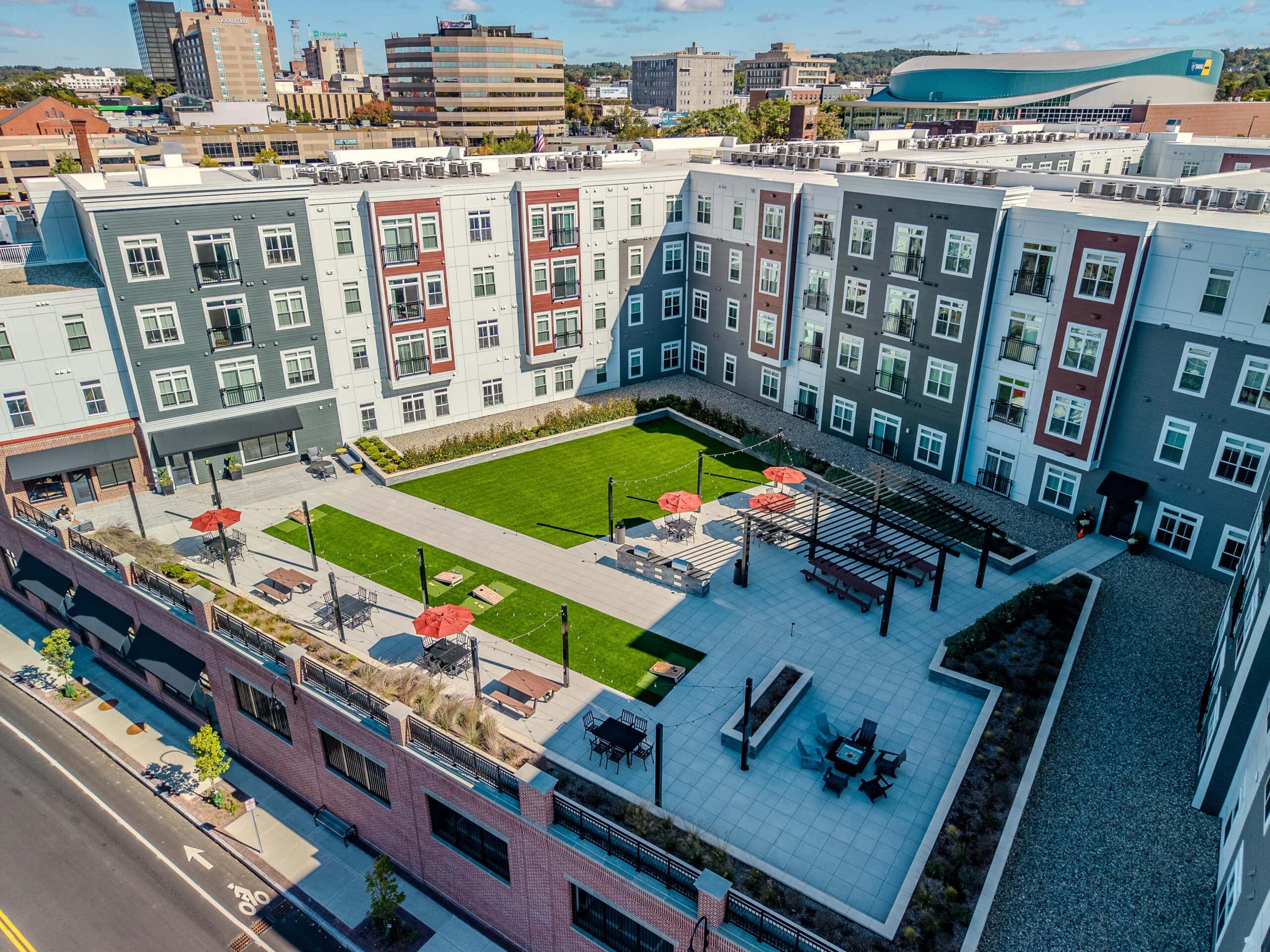 Drone view of the second floor courtyard amenity area with picnic tables shaded by pergolas. Artificial turf in the background. 75 Canal Apartments in Manchester, NH. Hardscaping and landscaping by Dex by Terra of Hudson, MA.