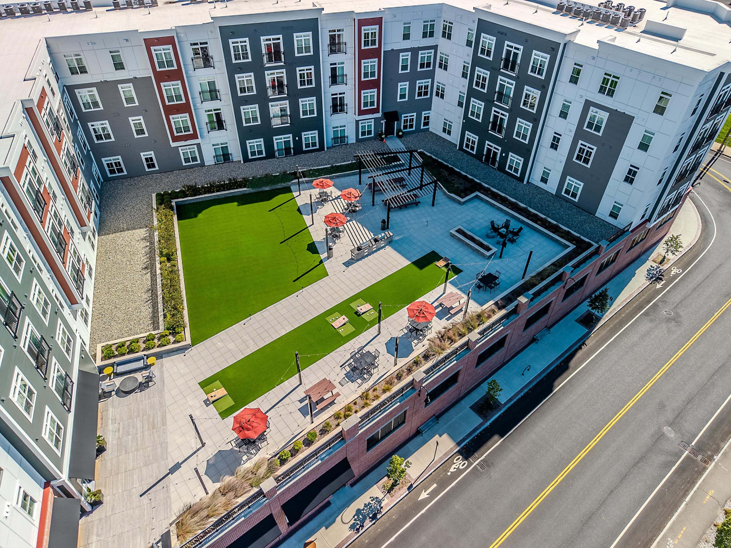 Drone view of the second floor courtyard amenity area with picnic tables shaded by pergolas. Artificial turf in the background. 75 Canal Apartments in Manchester, NH. Hardscaping and landscaping by Dex by Terra of Hudson, MA.