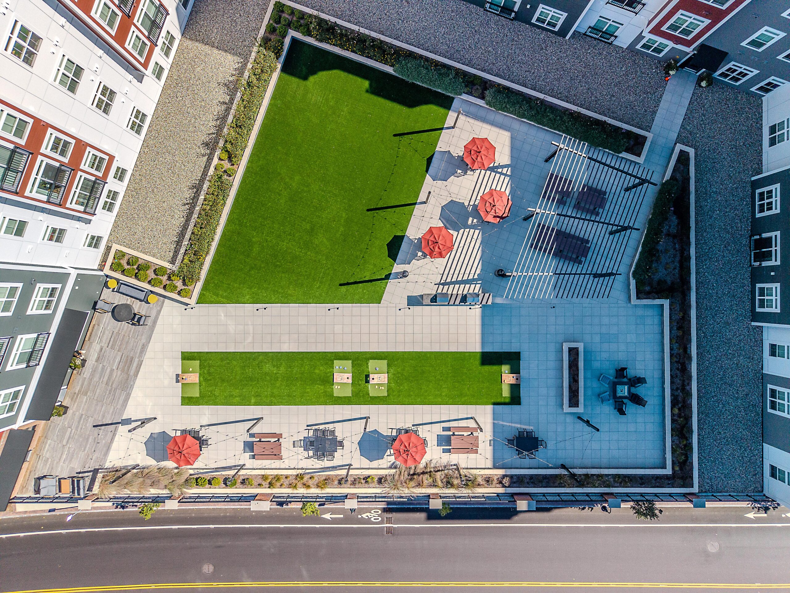Drone view of the second floor courtyard amenity area with picnic tables shaded by pergolas. Artificial turf in the background. 75 Canal Apartments in Manchester, NH. Hardscaping and landscaping by Dex by Terra of Hudson, MA.
