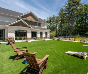 An artificial turf area for corn hole and other activities, with the clubhouse in the background at The Penstone Apartments in Walpole, MA. Hardscaping and landscaping by Dex by Terra of Hudson, MA.